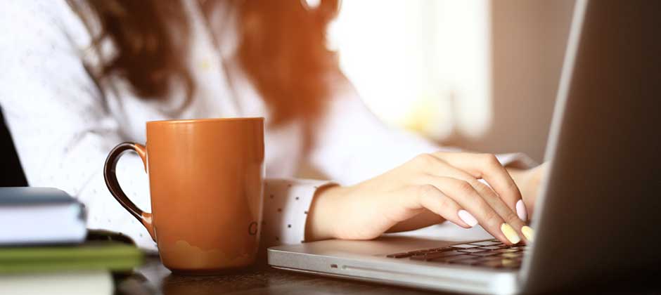 Person sitter med en kaffekopp å skriver på laptop